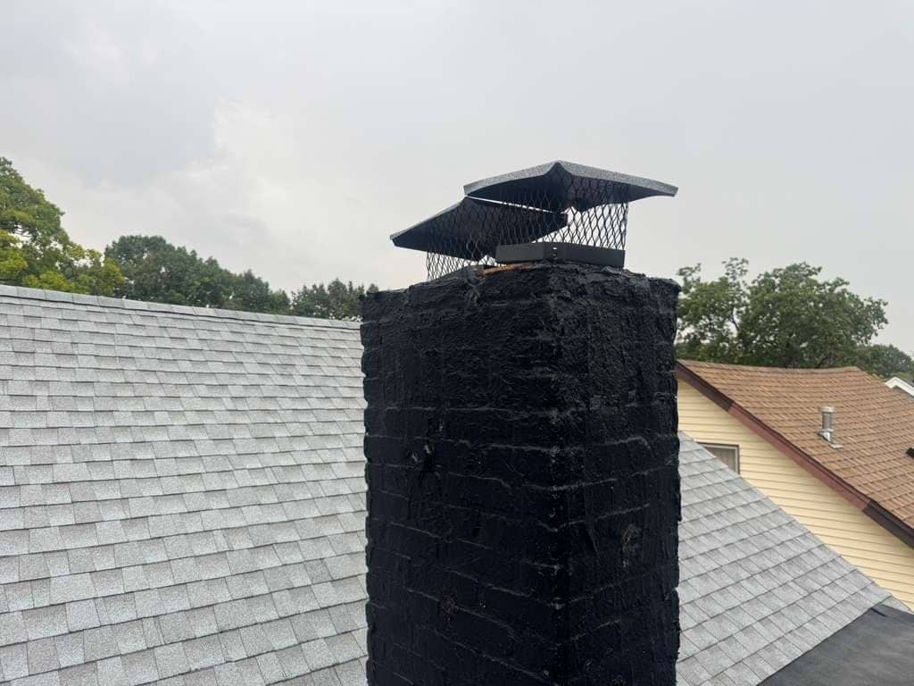 A black mesh chimney cap on top of a brick chimney coated in black, between shingle roofs under a grey sky.
