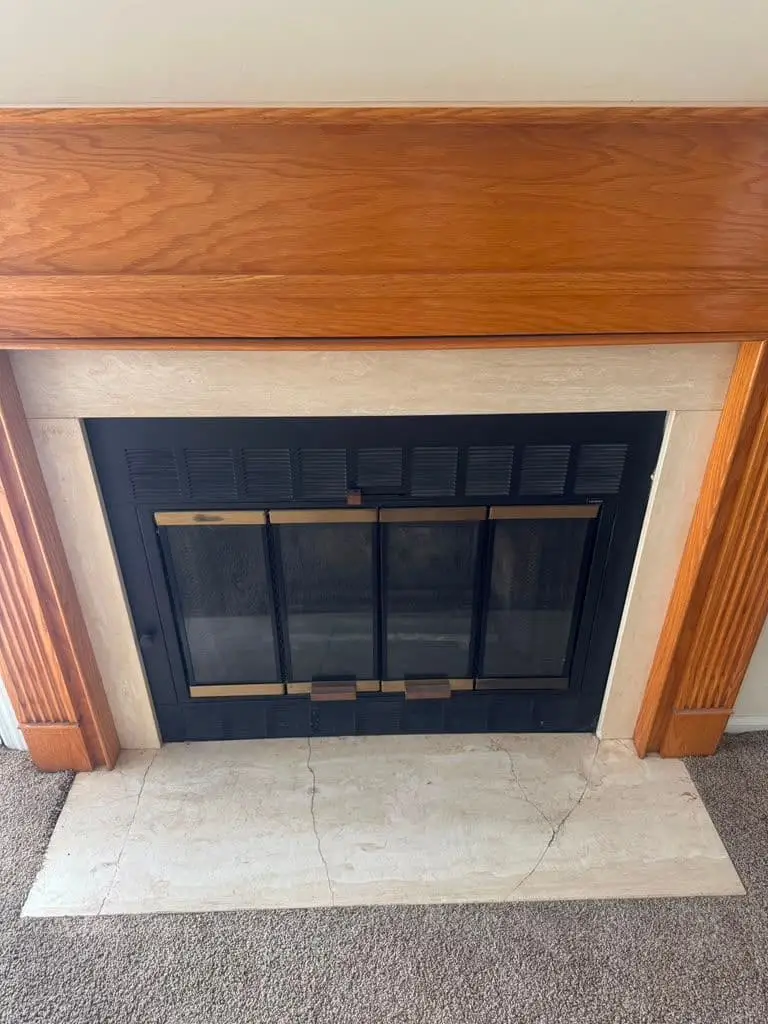 A glass fireplace door with brass trim and a louvered black frame under an oak mantel, above a cracked light marble hearth.