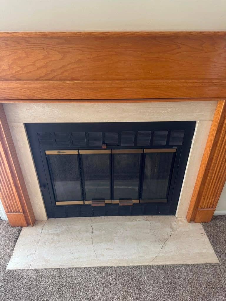 A glass fireplace door with brass trim and a louvered black frame under an oak mantel, above a cracked light marble hearth.
