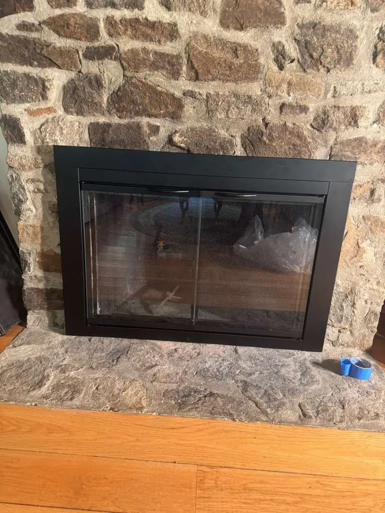A black-framed glass fireplace door with mesh screens fitted to a fieldstone fireplace, a roll of blue tape on the stone hearth.
